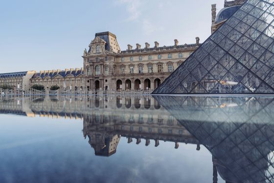 The Louvre Museum with its historic facade and glass pyramid reflected in a calm water surface under a clear blue sky.