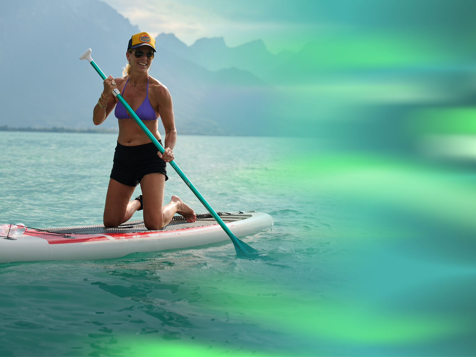 Woman kneeling on a paddleboard in a lake, wearing a cap, sunglasses, and a bikini top, with mountains in the background.