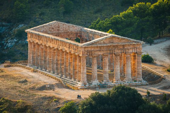 Ancient Greek-style stone temple with tall columns, set in a hilly landscape with surrounding trees and sunlight casting shadows.
