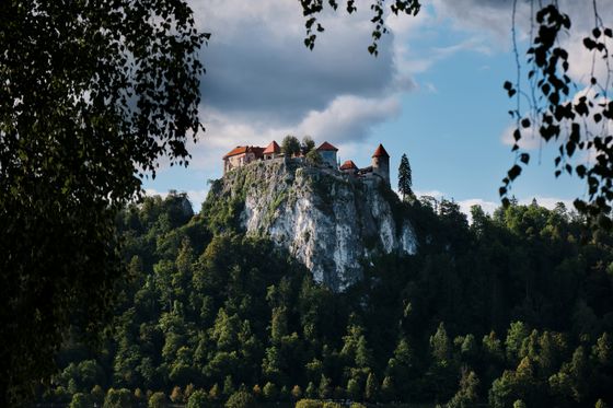 A castle with red roofs sits atop a rocky hill, surrounded by lush green trees, under a partly cloudy sky.