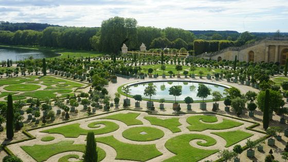 Ornate garden with geometric hedges, circular pond, and symmetrical pathways, surrounded by lush trees under a partly cloudy sky.