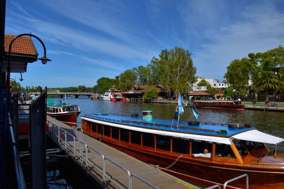 Wooden tour boats docked along a scenic river, with lush trees and buildings in the background under a clear blue sky.