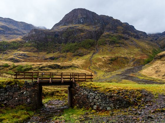 A wooden bridge over a stone wall with a path leading to a grassy mountain under an overcast sky.