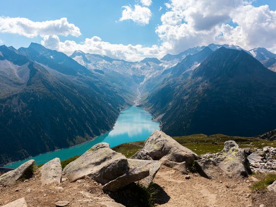 Scenic view of a turquoise lake nestled between rugged mountains under a partly cloudy sky, with rocky foreground.