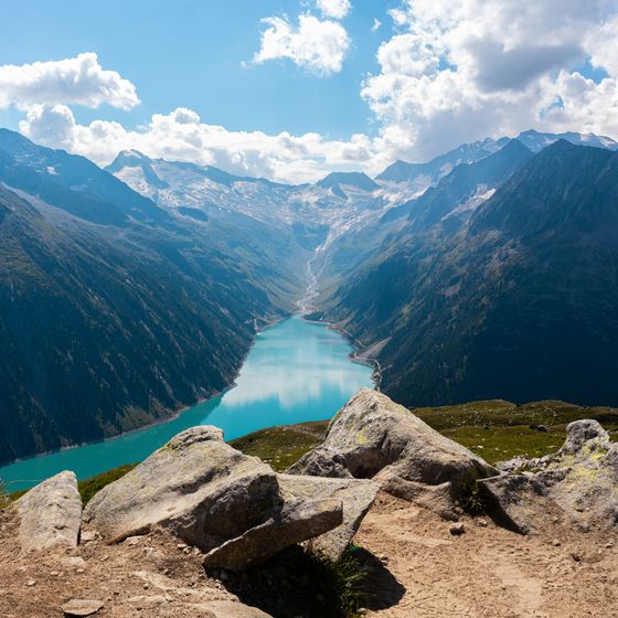 Mountain landscape with a turquoise lake winding through a valley, surrounded by rocky peaks under a partly cloudy sky.