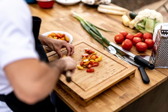 Person chopping tomatoes on a wooden cutting board, surrounded by fresh vegetables and kitchen utensils on a wooden table.