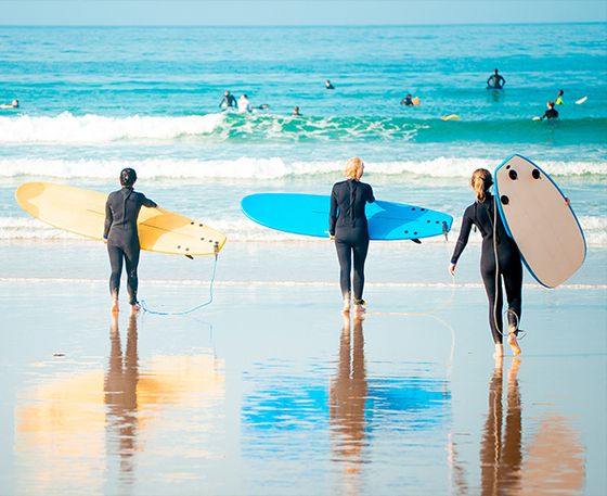 Three surfers in wetsuits carry surfboards toward the ocean. The beach is sunny, with other surfers visible in the water.