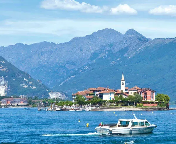 A boat sails on a lake with an island village and church, surrounded by mountains under a blue sky.