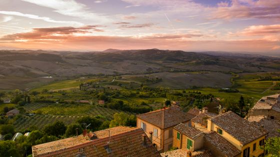 Scenic view of a Tuscan landscape at sunset, featuring rolling hills, vineyards, and rustic rooftops under a colorful sky.