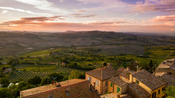 Scenic view of a Tuscan landscape at sunset, featuring rolling hills, vineyards, and rustic rooftops under a colorful sky.