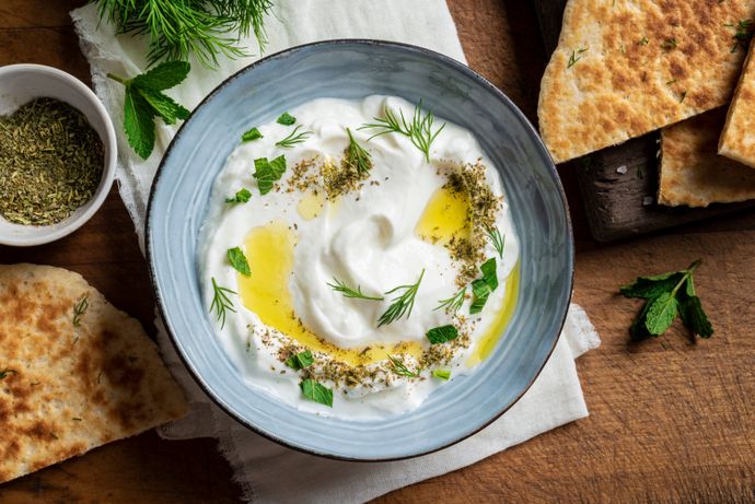 A bowl of creamy yogurt dip garnished with olive oil, herbs, and spices, surrounded by pita bread on a wooden surface.