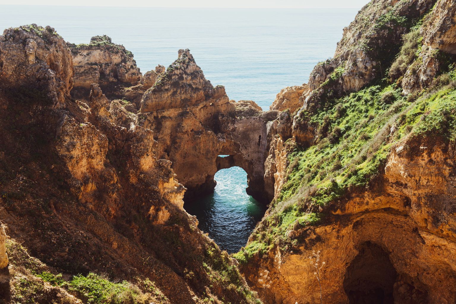 Rocky coastal cliffs with natural arches and a view of the ocean through the formations, surrounded by greenery.