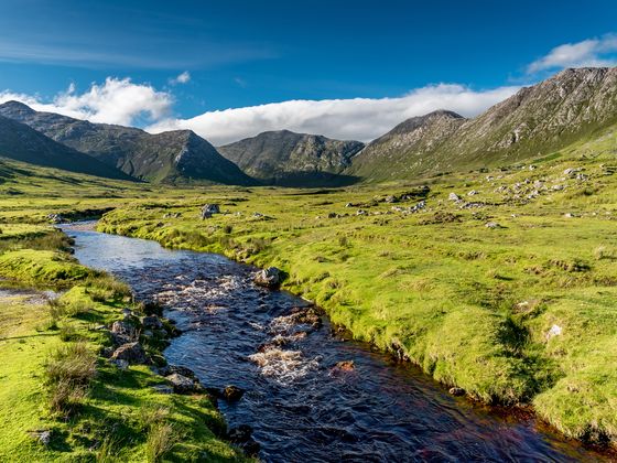 A serene landscape with a flowing river, lush green fields, and majestic mountains under a blue sky with scattered clouds.