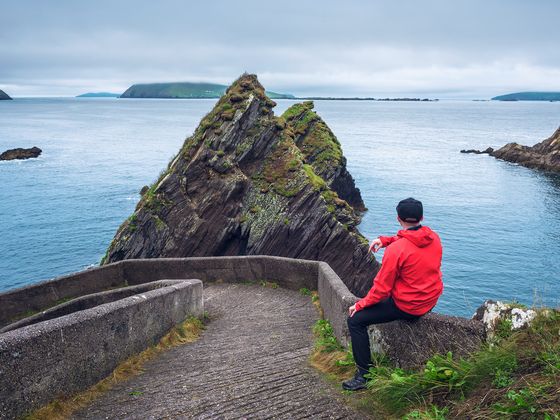 Person in a red jacket sits on a stone path, overlooking a rocky, moss-covered island in a calm blue sea under a cloudy sky.