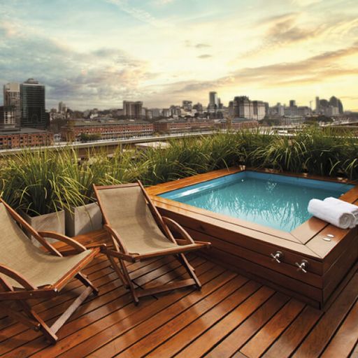 Rooftop deck with a small pool, two wooden lounge chairs, and city skyline view at sunset, surrounded by greenery.