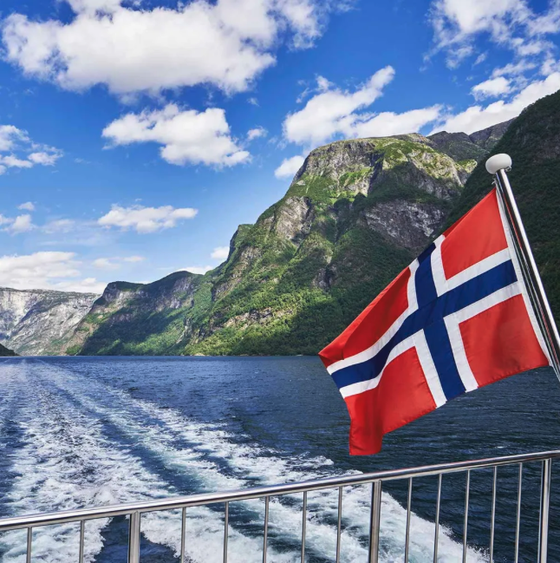 Norwegian flag on a boat with a wake trail, surrounded by mountains and a clear blue sky.