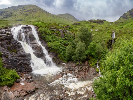 A picturesque waterfall cascades down rocky terrain surrounded by lush green hills and trees under a cloudy sky.
