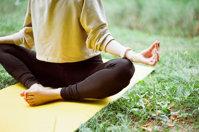 Person meditating in a cross-legged position on a yellow yoga mat outdoors, surrounded by grass.