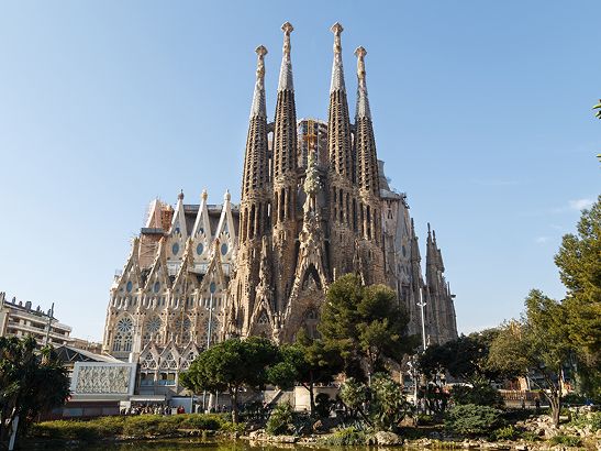 Gothic-style basilica with tall spires and intricate facades, set against a clear blue sky, surrounded by trees and a reflective pond.