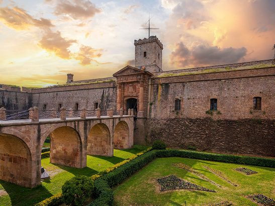 Stone fortress with arched bridge, green manicured gardens, and a dramatic sunset sky.