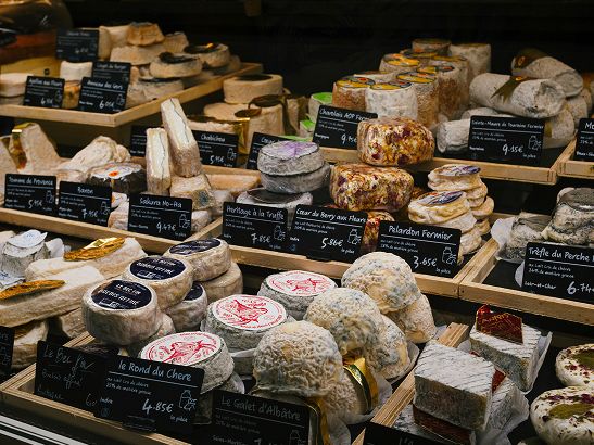An assortment of cheeses displayed on wooden shelves, each with a label indicating name and price, showcasing various textures and shapes.