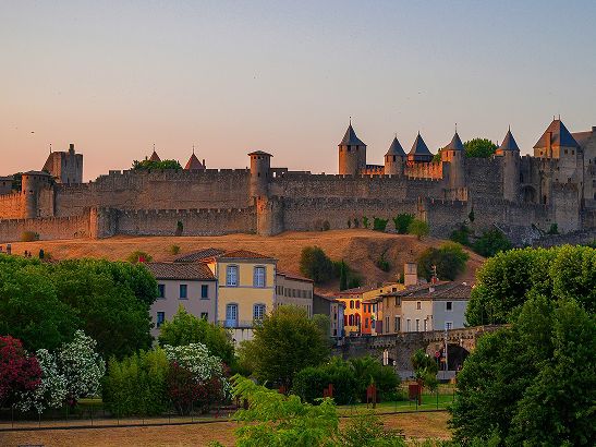 View of the medieval fortress of Carcassonne at sunset, with the fortified walls and towers overlooking surrounding houses and lush greenery.