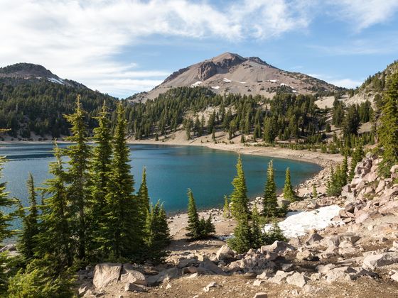 A serene mountain lake surrounded by pine trees and rocky terrain, with a distant peak under a partly cloudy sky.