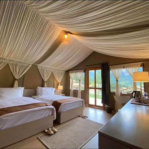 Luxurious tent interior with two beds, draped ceiling, and a view of greenery through large windows. Cozy ambiance with warm lighting.