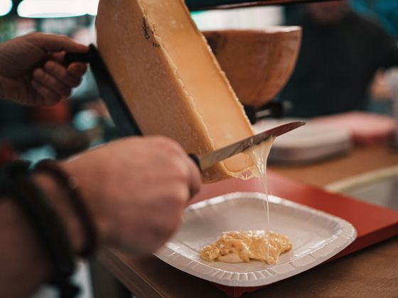 A person scrapes melted cheese onto a plate with a knife, serving a creamy dish in a cozy indoor setting.