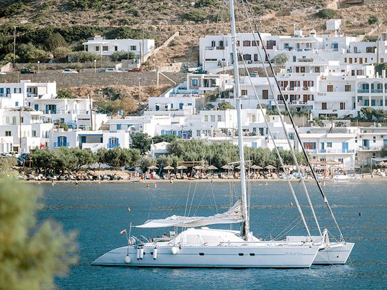A sailboat anchored in blue waters with a backdrop of white hillside buildings and greenery under a clear sky.