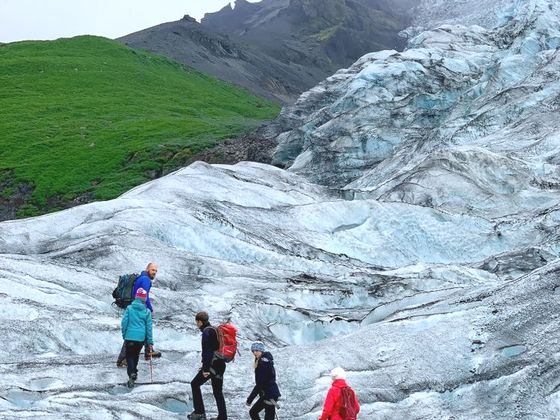 Five hikers with backpacks trek across a glacier with rocky mountains and a green hill in the background under a cloudy sky.