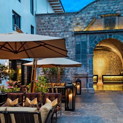 Outdoor hotel courtyard with seating, umbrellas, lanterns, and stone archway. Evening ambiance with warm lighting and lush plants.