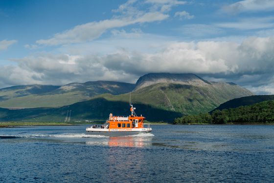 Orange ferry boat on a calm lake with passengers, surrounded by lush green hills and a partly cloudy sky.