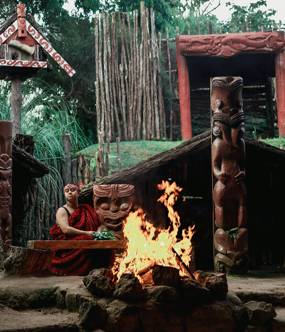 A person in traditional attire sits by a fire, surrounded by wooden carvings and structures, in a lush outdoor setting.