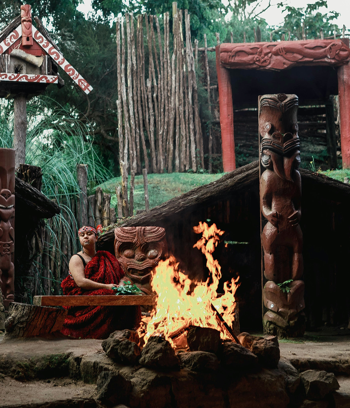 A person in traditional attire sits by a fire, surrounded by wooden carvings and structures, in a lush outdoor setting.