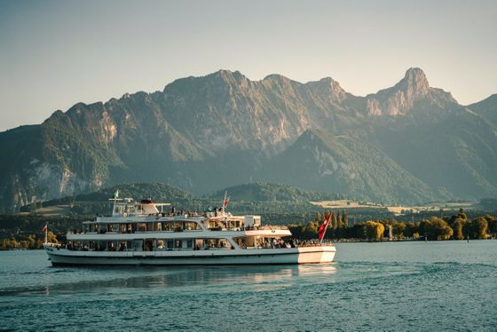 A large white ferry with passengers sails on a calm lake, with majestic mountains and a clear sky in the background.