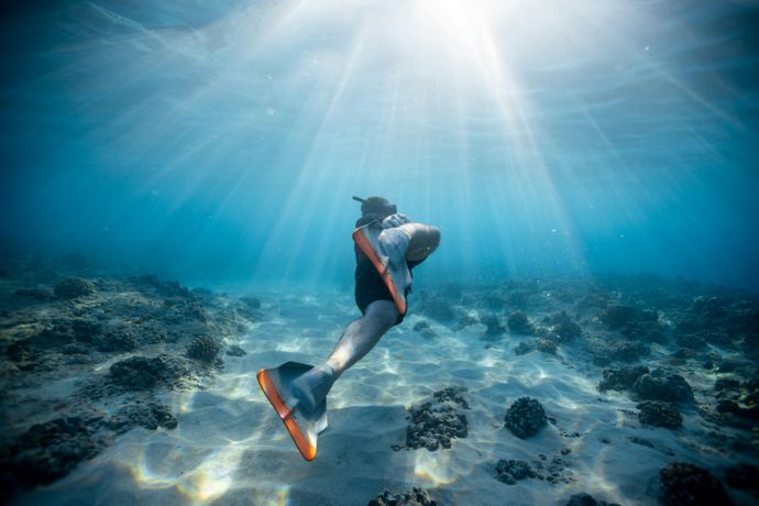 A snorkeler with fins swims underwater in clear blue ocean, sunlight streaming through, illuminating the sandy seabed and scattered coral.