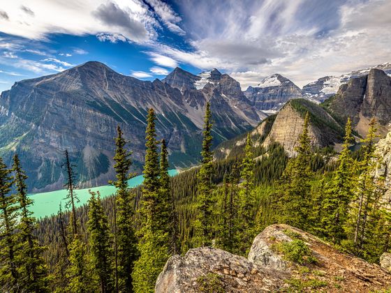 Majestic mountain range with a turquoise lake below, dense evergreen forest, and a dramatic cloudy sky.