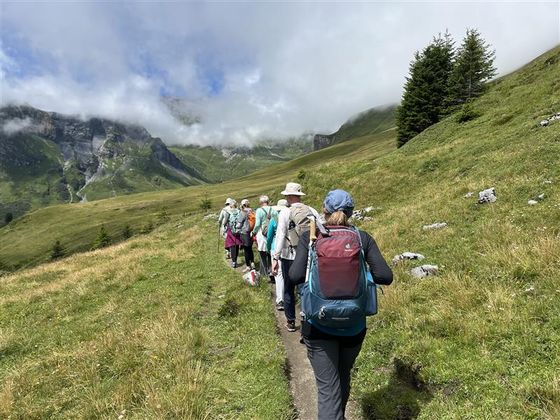 Hikers with backpacks walk along a narrow trail through a lush, green mountain landscape under a partly cloudy sky.