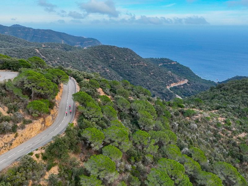 Aerial view of cyclists on a winding road through lush green hills, with the ocean in the background under a partly cloudy sky.