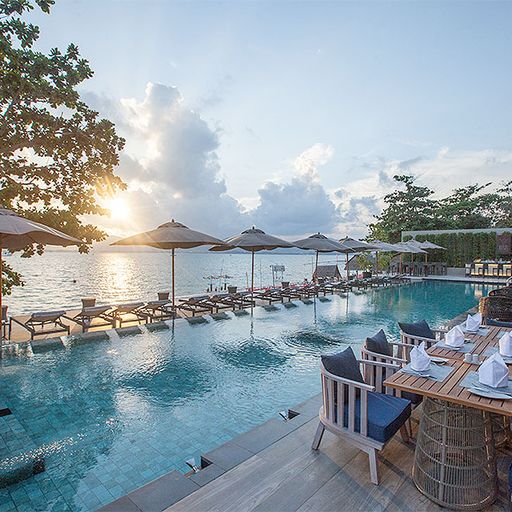 Infinity pool with sun loungers and umbrellas by the ocean at sunset, surrounded by trees and a dining area with neatly set tables.