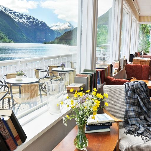 Cozy room with books, a vase of wildflowers, and a scenic view of a lake and mountains through large windows.
