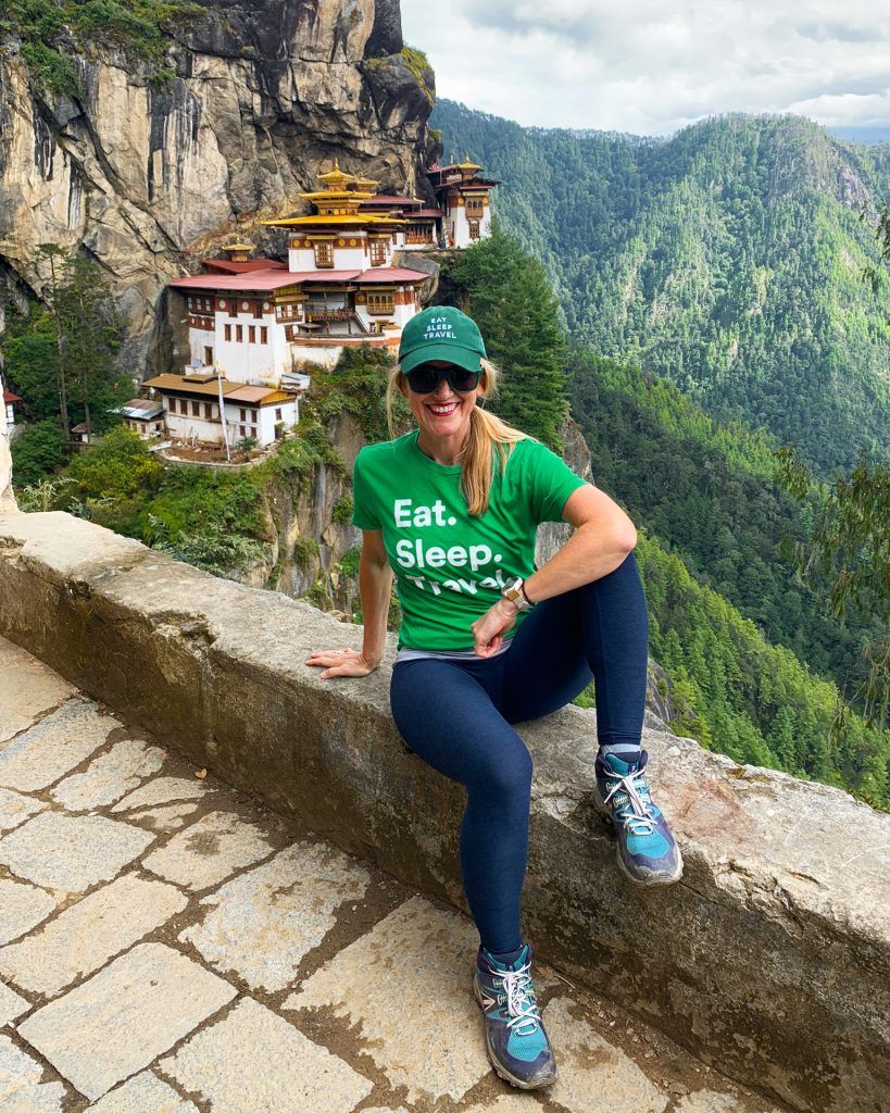 Woman in a green shirt and cap sits by a stone railing with a view of the Tiger's Nest Monastery in Bhutan, lush green mountains in the background.