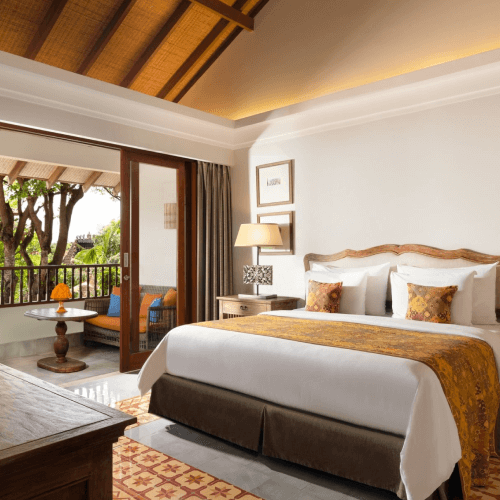 Elegant hotel room with a large bed, wooden furniture, and a balcony overlooking greenery. Warm lighting and decorative pillows enhance coziness.