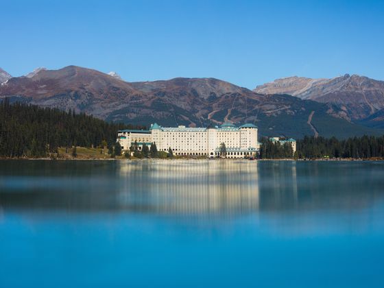 A large, elegant hotel stands by the serene turquoise lake, surrounded by evergreen forests and majestic mountains under a clear blue sky.