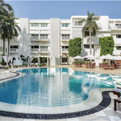 Outdoor hotel pool with surrounding lounge chairs, cabanas, and palm trees. White building in the background under a clear blue sky.