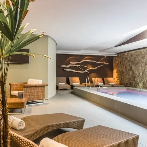 Modern indoor spa with wicker loungers, a rectangular pool, stone accent wall, and tropical plants, creating a serene and relaxing atmosphere.