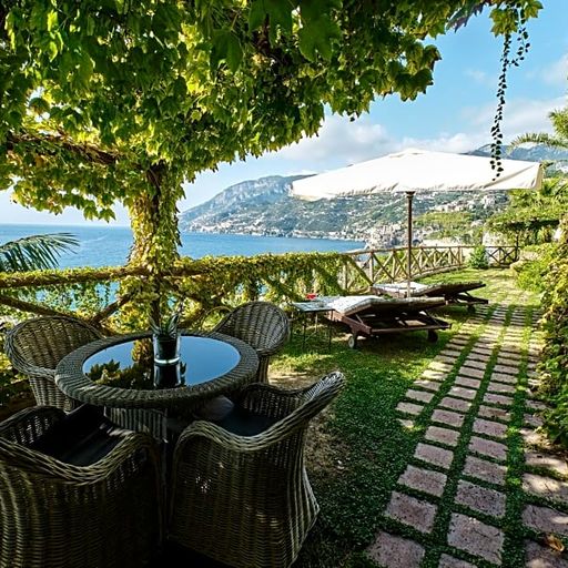 Outdoor patio with wicker chairs around a glass table, shaded by a tree. Overlooks a scenic coastal view with mountains and sea.