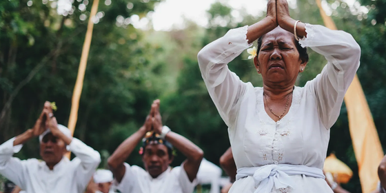 People in traditional white attire perform a prayer ritual outdoors, with hands pressed together, set against a lush green background.
