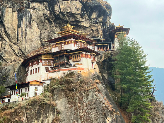 A monastery perched on a cliffside, surrounded by trees and rugged rock, under an overcast sky.
